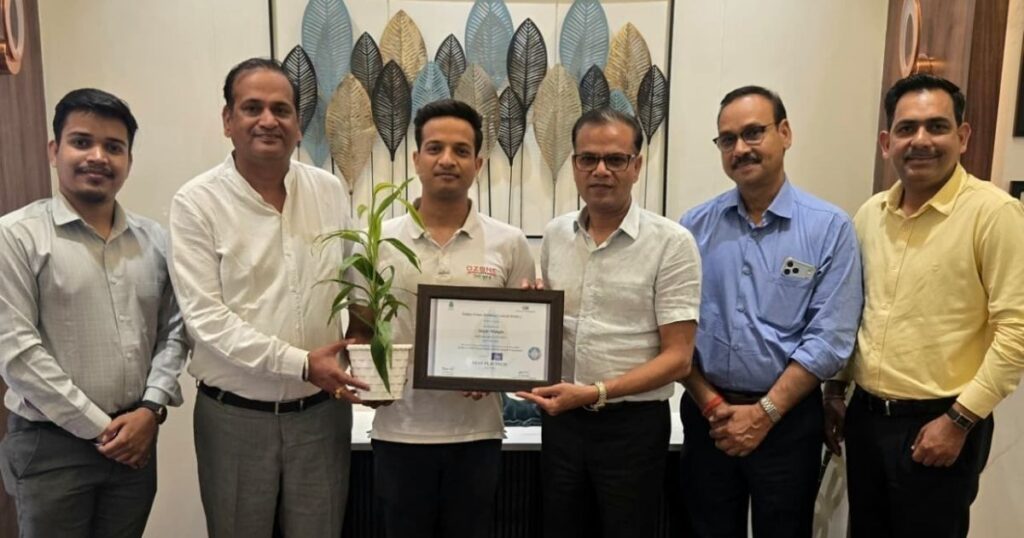 Six men stand indoors; one holds a potted plant while another holds a framed certificate, signaling an award presentation or recognition event.